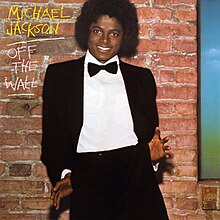 A smiling male (Michael Jackson) with a black afro, wearing a black tuxedo, white shirt, and a black bow tie. Both of his thumbs are hooked into his pants pockets with his palms and fingers facing forward and splayed out. The sides of his jacket are tucked behind his hands as he leans back slightly, giving a playful, casual touch to his formal look. Behind him there is a brown brick wall and to the side of his head are MICHAEL JACKSON in yellow chalk writing and OFF THE WALL in white chalk writing. JACKSON and WALL are separately underlined.