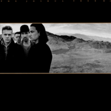 A landscape monochrome photograph of U2 in the desert sits in the center of a black background. U2 are standing on the left half of the photograph, with a mountain range on the right half. Tiny gold text reading THE JOSHUA TREE U2 is stretched across the top of the black background.